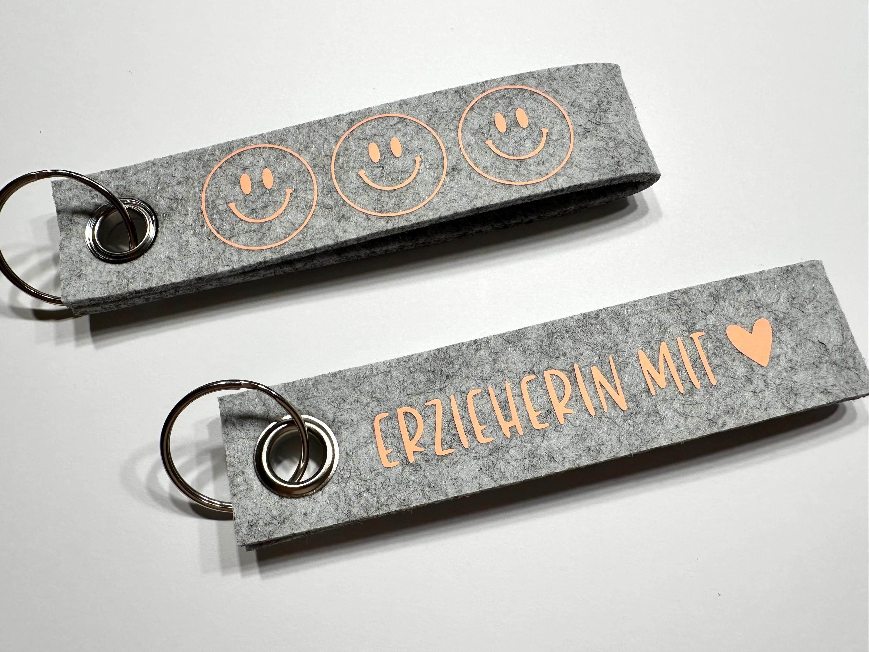 Zwei Schlüsselanhänger aus grauem Filz. Einer zeigt drei lächelnde Gesichter in Pastell Orange . Der andere hat den Text "ERZIEHERIN MIT" und ein orangefarbenes Herz. Beide haben silberne Schlüsselringe.