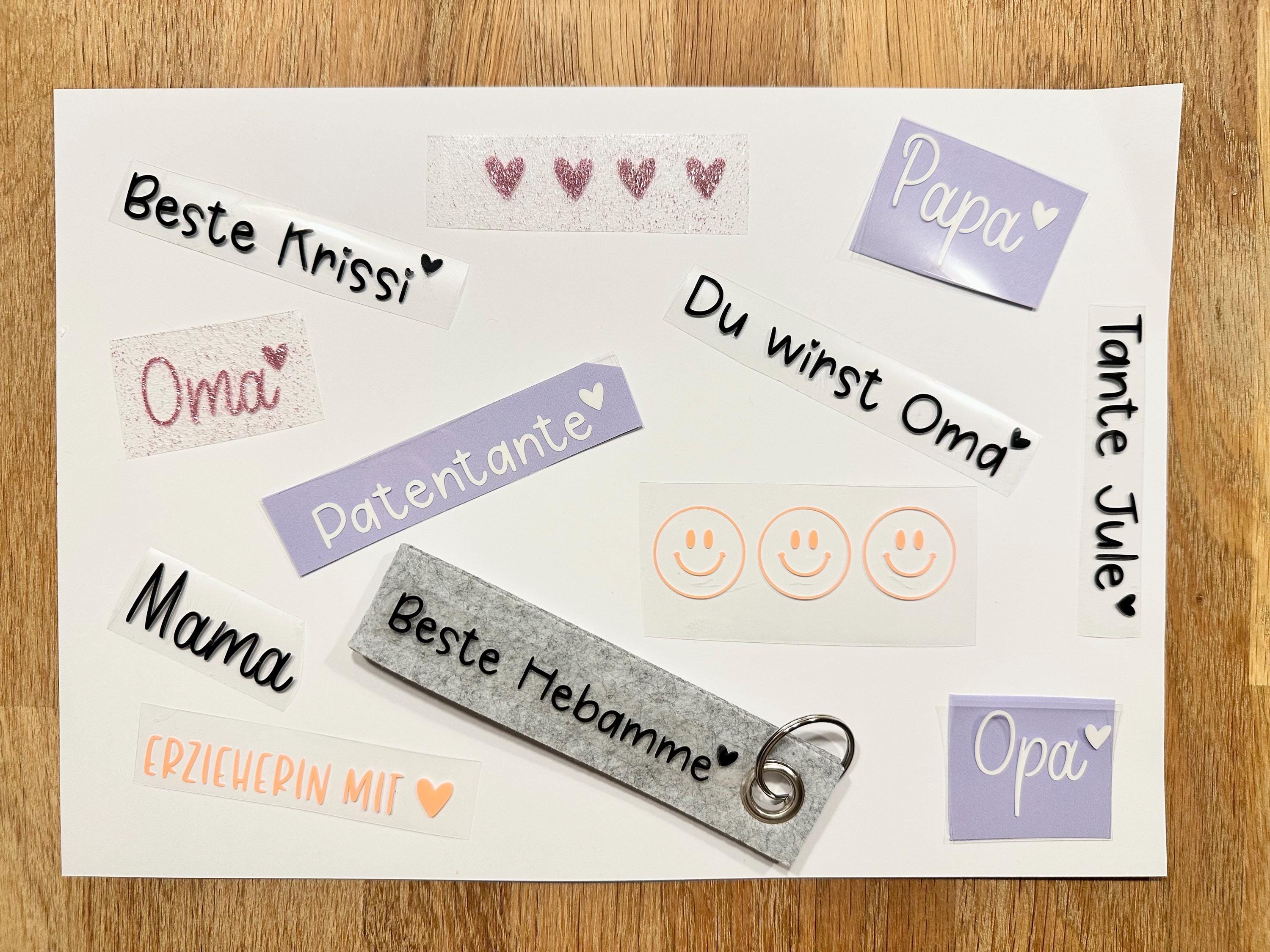 Eine Sammlung von Aufklebern und einem Schlüsselanhänger mit verschiedenen deutschen Sätzen. Die Aufkleber enthalten Sätze wie "Mama", "Oma" und "Papa" sowie Smiley-Gesichter und Herz-Akzente.