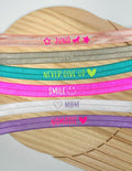 Eine Auswahl an elastischen Armbändern in verschiedenen Farben, jeweils mit einem anderen Wort oder Satz bedruckt. Die Bänder enthalten die Wörter "Juna", "Happy", "Never Give Up", "Smile", "Mom" und "Namaste".