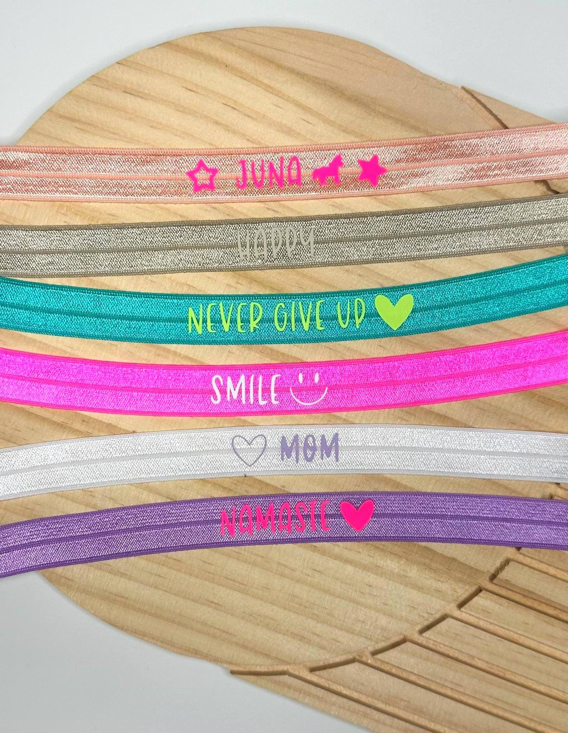Eine Auswahl an elastischen Armbändern in verschiedenen Farben, jeweils mit einem anderen Wort oder Satz bedruckt. Die Bänder enthalten die Wörter "Juna", "Happy", "Never Give Up", "Smile", "Mom" und "Namaste".